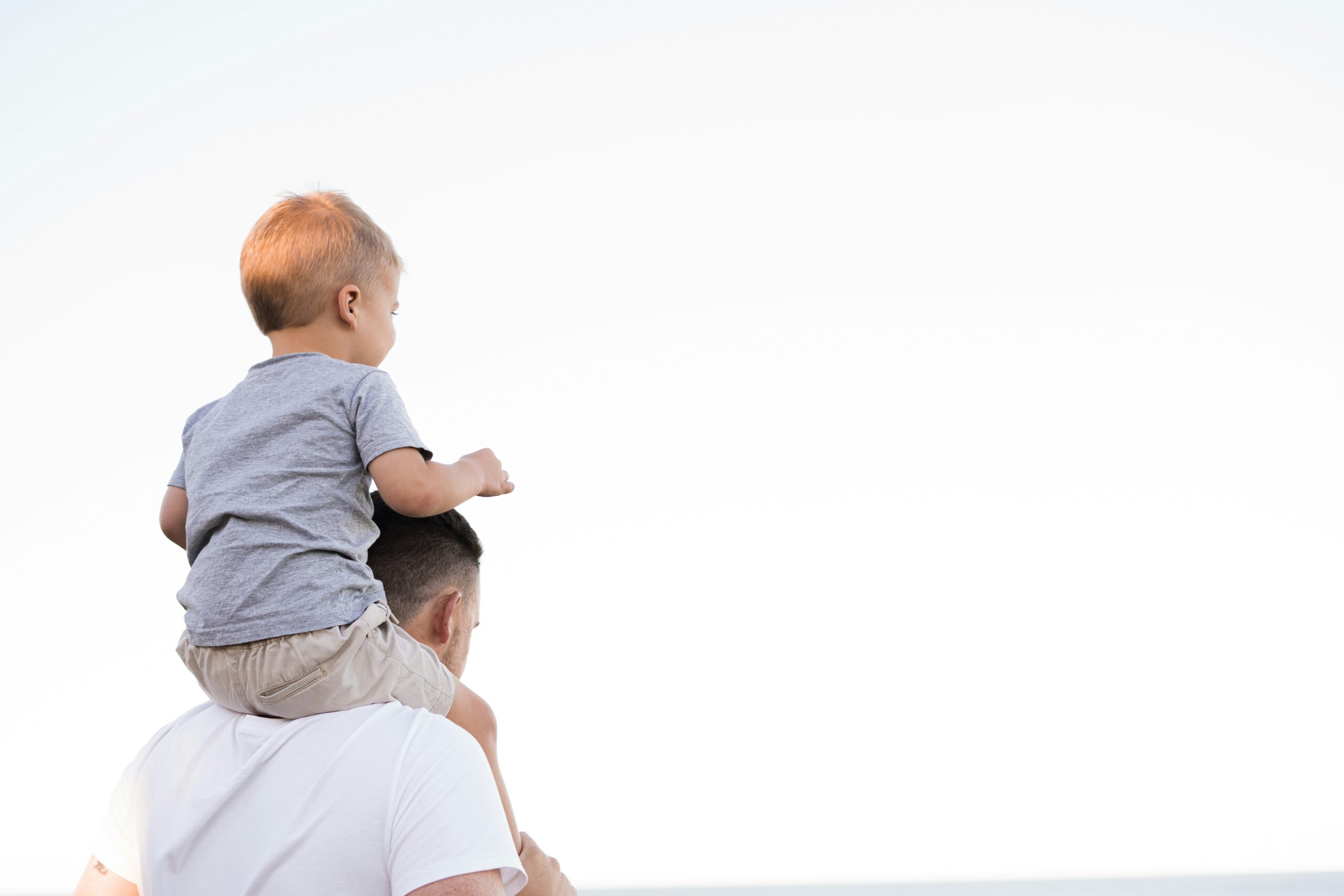 Vater steht mit dem Rücken zu uns und blickt in die Ferne. Auf den Schultern sitzt sein Sohn.